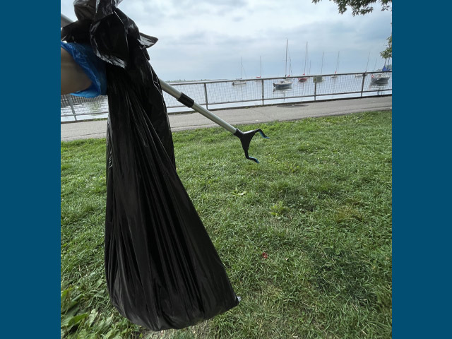 A person holding a trash bag and grabber at a grassy park overlooking water.