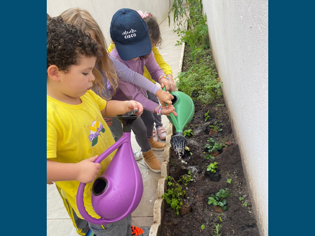 Four children holding watering cans watering plants in a small garden.