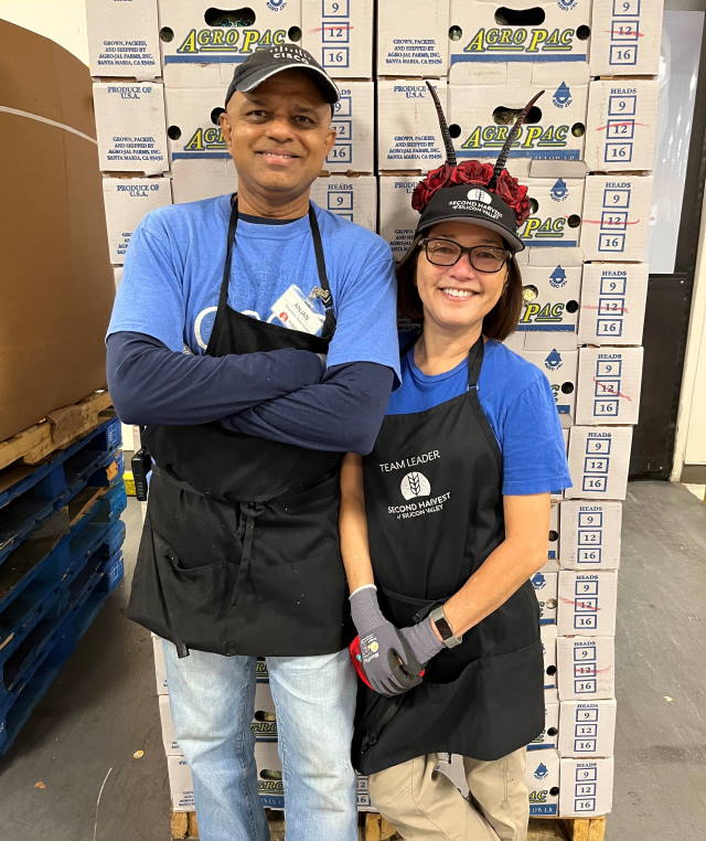 Anjan and Amy smiling and  standing in front of a large stack of boxes at Second Harvest.