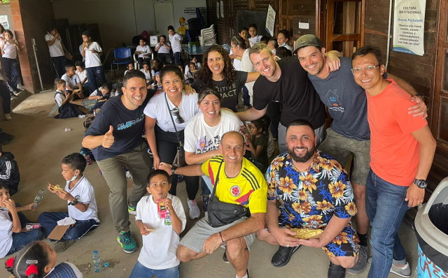 A large group of people pose with  children at a school in Colombia while eating lunch.