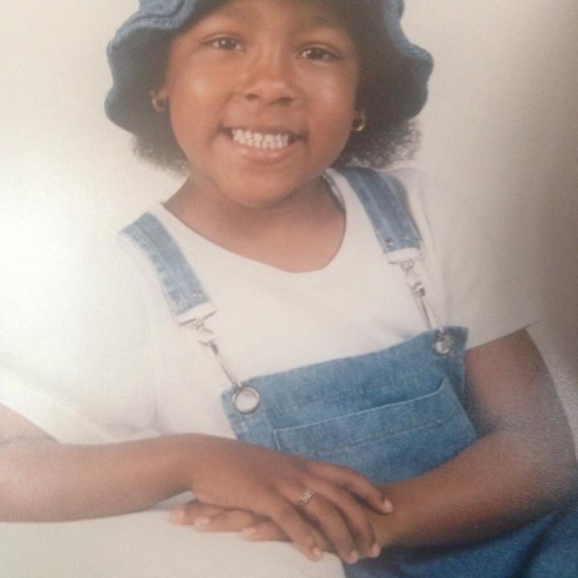 Young Andrea smiles, wearing denim overalls and hat.