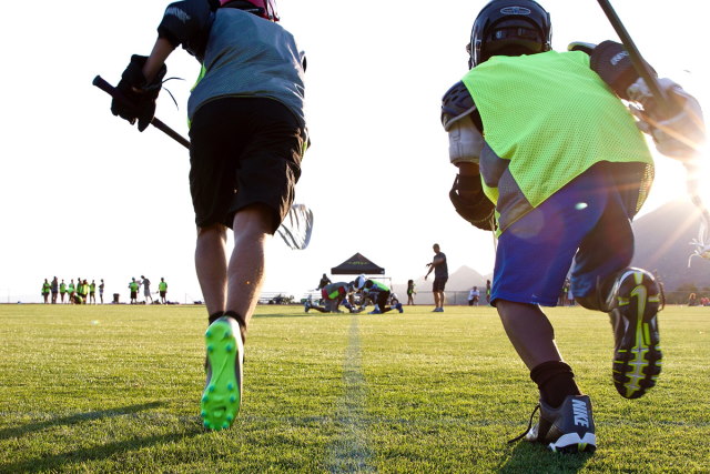 Two children running on a field wearing  lacrosse gear.