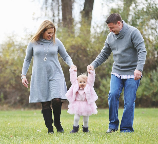 Kelly and Alan holding Lyla’s hands in a field of grass and bushes.