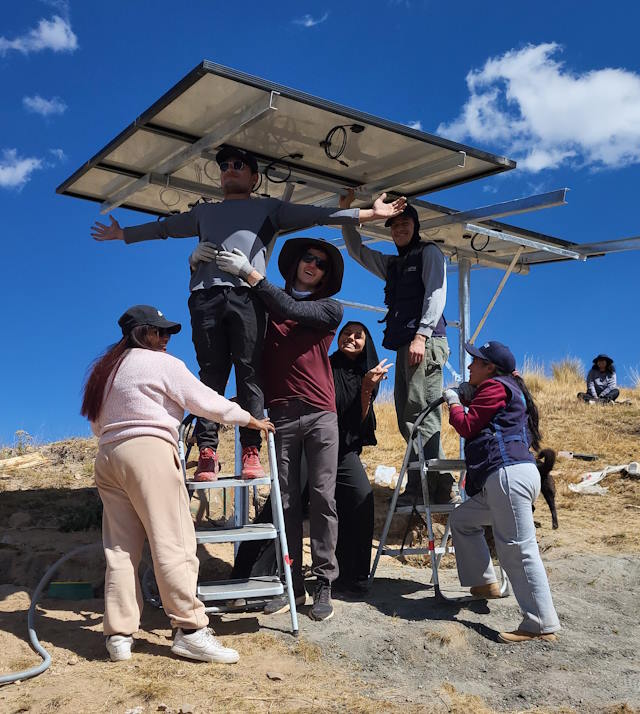 A group of smiling people installing a solar panel on a sunny hill.