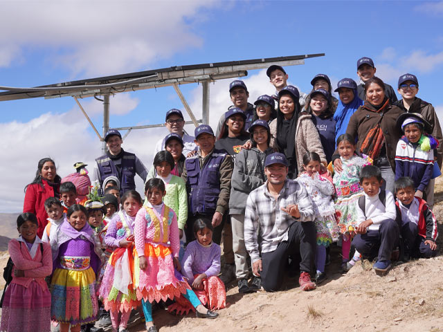 Children in Andean clothing and adults in volunteer hats stand near solar panels.