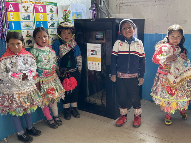 Five children in colorful clothes stand beside a large mechanical box in a classroom.