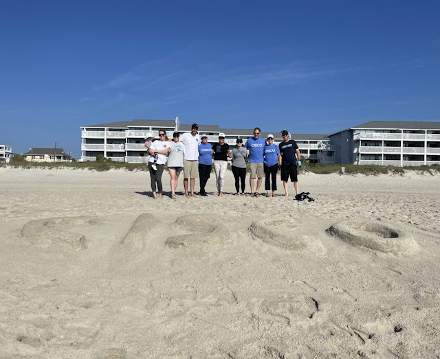A small group of people standing on a beach behind “Cisco” spelled out in the sand.
