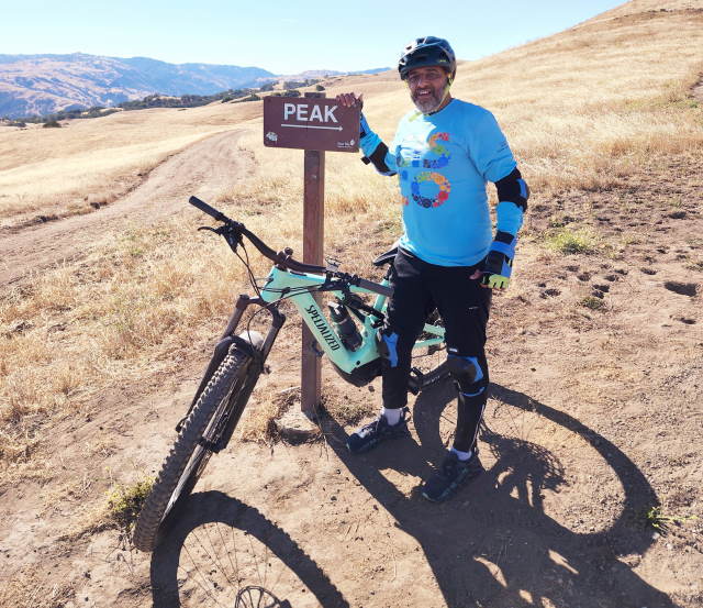 Rohan Naggi stands with his bike in front of a  sign pointing to the mountain peak.