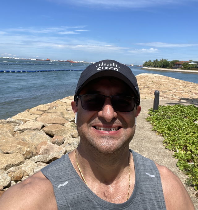 Sanjay Kaul smiles in a Cisco hat while outside near a large body of water.