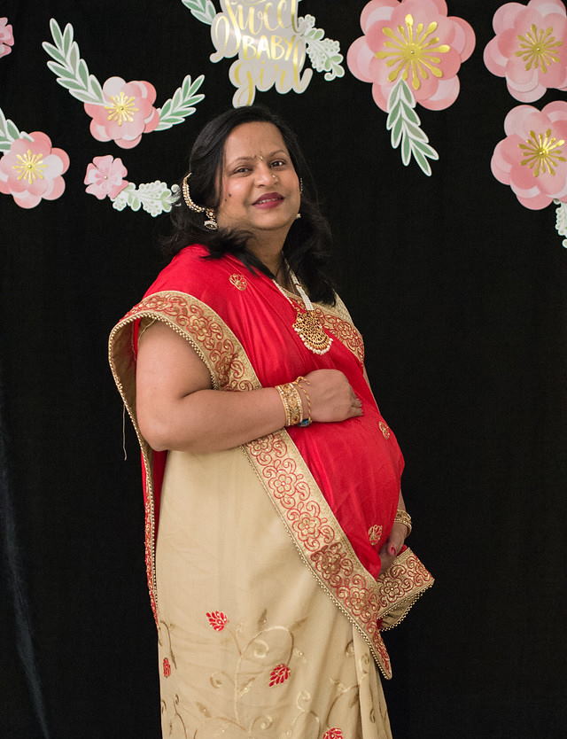 A pregnant Hetal standing against  a black background with pink flowers.