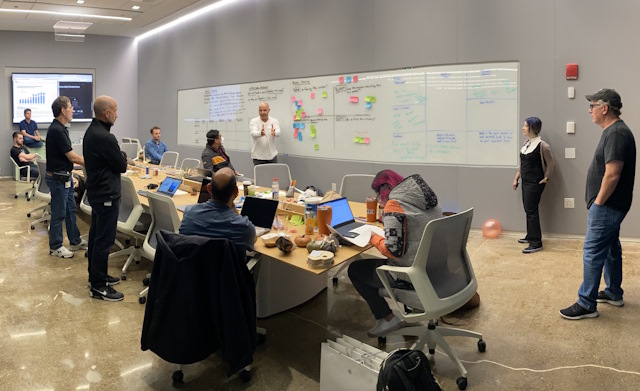 A team collaborating in a meeting room around a whiteboard with text and sticky notes.
