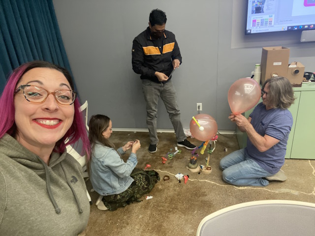 A woman holding the camera while three team members build with balloons and other items.