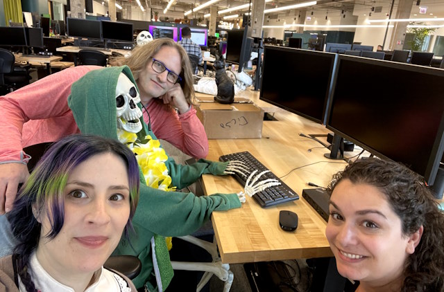 Three people pose with a skeleton seated at a workstation, dressed in a green hoodie and yellow flower lei.